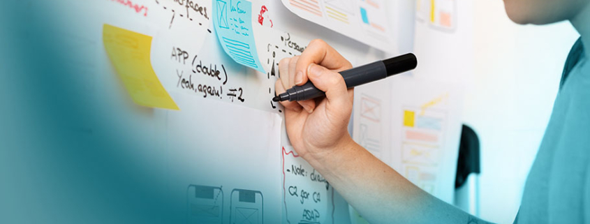 Hybrides Projektmanagement: Person schreibt auf Whiteboard mit Notizen und Diagrammen. Komplexe Projektplanung sichtbar durch Formeln, Skizzen und farbige Post-its.