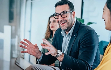 Ein Mann mit Brille und Sakko gestikuliert und lächelt in einem Meeting, während er mit zwei Frauen in einem modernen Büro spricht. Grundlagen Change Management