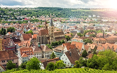 Stuttgart: Luftaufnahme Stuttgarts mit historischer Altstadt, roten Ziegeldächern und grünen Hügeln bei warmem Sonnenlicht.