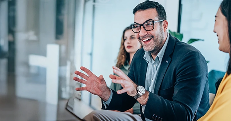 Ein Mann mit Brille und Sakko gestikuliert und lächelt in einem Meeting, während er mit zwei Frauen in einem modernen Büro spricht. Grundlagen Change Management