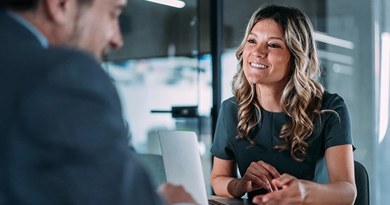 Frau mit lockigem Haar im Gespräch mit Mann am Schreibtisch, beide in modernem Büro, konzentrierte Gestik und freundliche Mimik. Erfolgreiche Verhandlungsführung im Einkauf