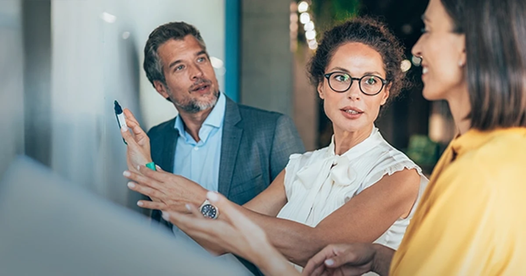 Gespräch am Whiteboard, Frau in Weiß zeigt etwas, Kollege hört zu, unscharfer Vordergrund links, moderne Büroumgebung. Führen mit Zielen