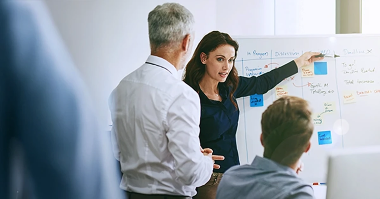 Geschäftsfrau in dunkler Business-Kleidung erklärt enthusiastisch vor Whiteboard mit farbigen Post-its, Kollegen hören zu. Führen von Führungskräften