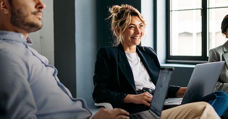 Fröhliche Geschäftsfrau mit Laptop in hellem Büroraum, Team-Kollegen arbeiten im Hintergrund. Grundlagen der Führung