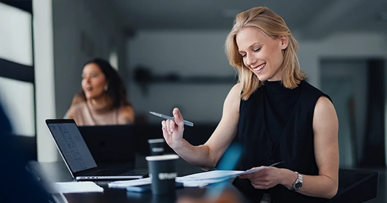 Blonde Frau arbeitet lächelnd mit Stift und Papier am Tisch, Laptop daneben, Kollegin unscharf im Hintergrund. Agiles Projektmanagement