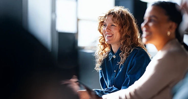 Frau mit Locken lacht in einer Gruppe, helle Büroumgebung mit weichem Licht und Sitzreihe. Grundlagen Projektmanagement