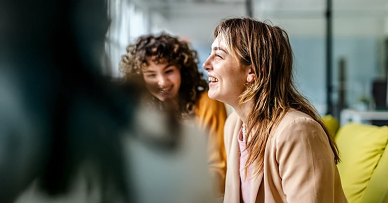 Zwei Frauen sitzen auf einem Sofa und lachen, unscharfe Person im Vordergrund, helles Büro mit Glaswänden. Hybrides Projektmanagement