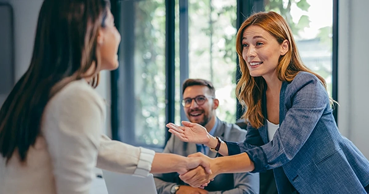Projektmanagement Seminar - Projektleiter Weiterbildung lernen Personen kennen Rothaarige Frau schüttelt einer Kollegin die Hand, Mann mit Brille sitzt lächelnd am Tisch, Stakeholdermanagement für den Projekterfolg