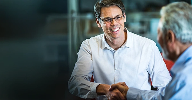 Handschlag zwischen zwei Männern am Tisch, Mann mit Brille lächelt, moderne Umgebung mit Glas und neutralen Farben. Strategische Kommunikation