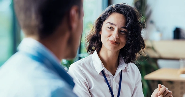 Junge Frau mit dunklen Haaren und weißer Bluse hört aufmerksam einem Mann zu, weiche Bürohintergründe mit Pflanzen und Fenstern. Key Account Management